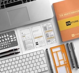 A neatly arranged workspace features a laptop, a UX sketchbook, a keyboard, drawing tools, stencil templates, and wireframe sketches for a mobile app design. The environment appears organized and intended for user experience design work.