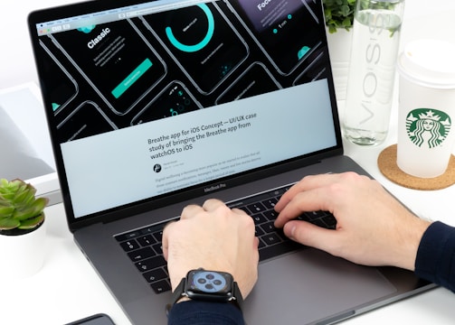 A person is typing on a MacBook Pro laptop surrounded by a smartphone, a green plant, a bottle of VOSS water, and a Starbucks paper cup. The laptop screen displays a UI/UX design with several circular graphic elements and a heading related to an iOS concept. The individual is wearing a smartwatch on their wrist.