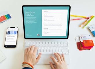 A person is using a tablet with a keyboard on a desk. The tablet displays a screen about search function design and considerations. Nearby, there is a smartphone showing some exploration and creation options. Various colorful cards and pens are scattered on the white surface.
