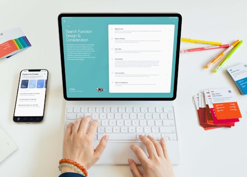 A person is using a tablet with a keyboard on a desk. The tablet displays a screen about search function design and considerations. Nearby, there is a smartphone showing some exploration and creation options. Various colorful cards and pens are scattered on the white surface.